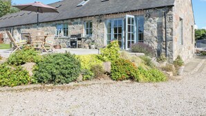 A stone building with a gravel driveway, a patio with outdoor furniture, and a large umbrella. - Stable Cottage (Inverurie)