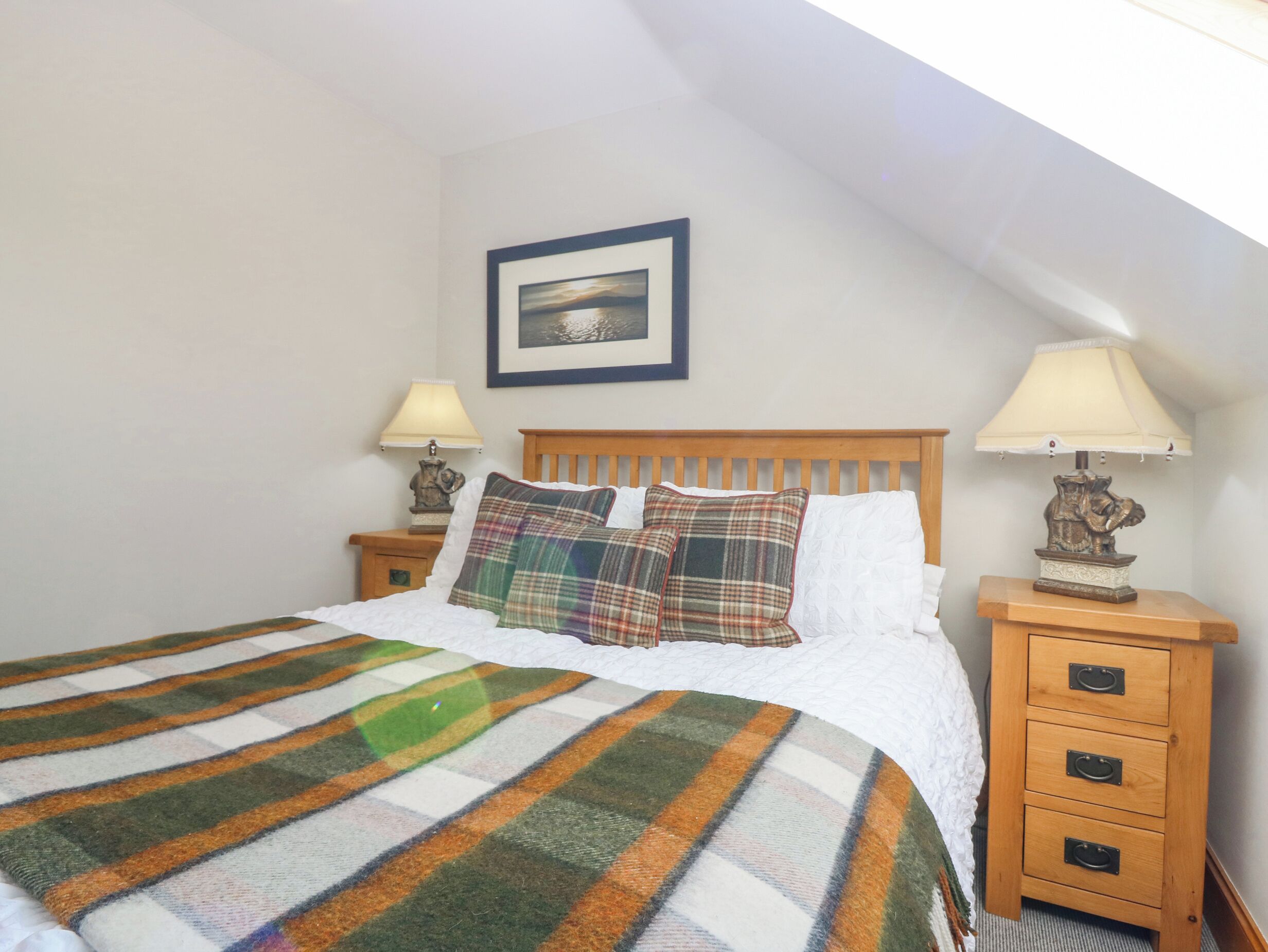 A neatly made bed with a striped blanket, two bedside tables with lamps, a framed picture on the wall, and a sloped ceiling.