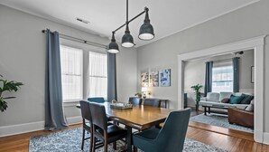 Un comedor con una mesa de madera, sillas e iluminación colgante. Se observa un sofá y una mesa auxiliar al fondo.