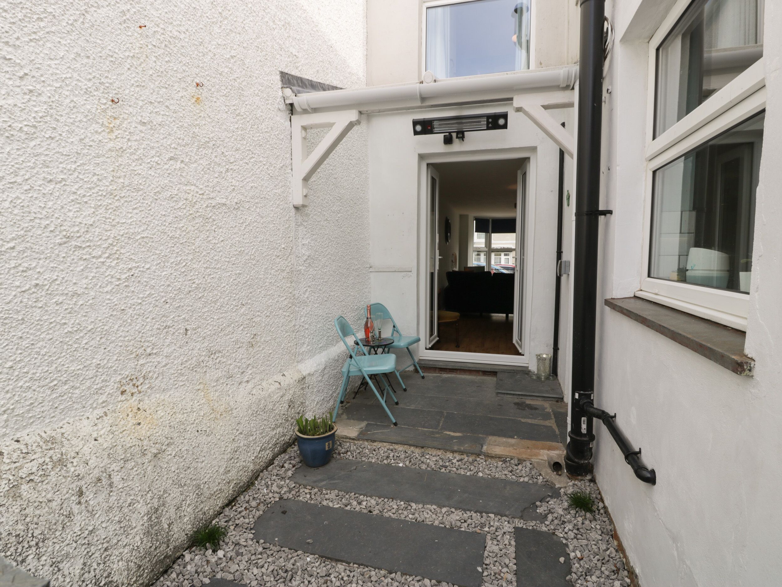 A narrow outdoor corridor with a white textured wall, a small table with chairs, and a potted plant.