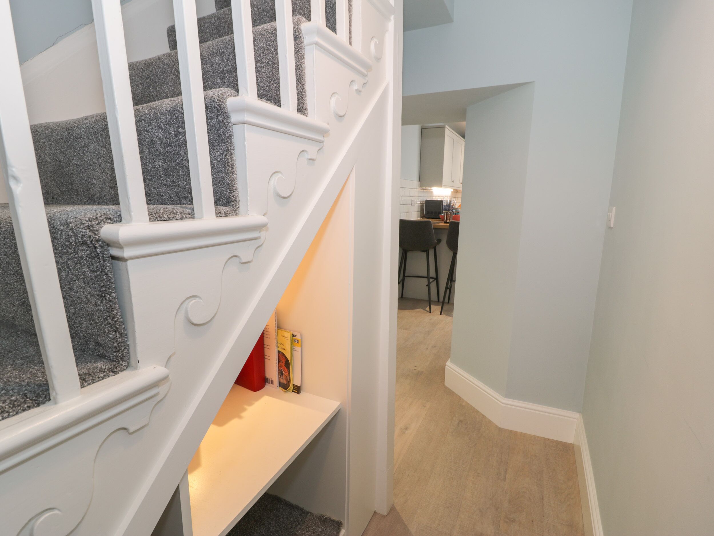 A staircase with a white handrail leading to a landing. Below the landing, there is a white shelf with a red object on it. The room has light blue walls and a wooden floor.