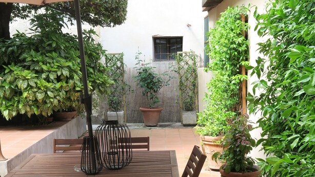 Eine Terrasse mit Holztisch und -stühlen, umgeben von Kübelpflanzen und einem großen Sonnenschirm.