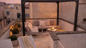 A rooftop terrace with lounge chairs, a sofa, and a small table, set against a sunset backdrop.