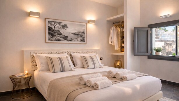 A bedroom with a large bed, bedside lamps, a framed picture, a wardrobe, and a window with a view.