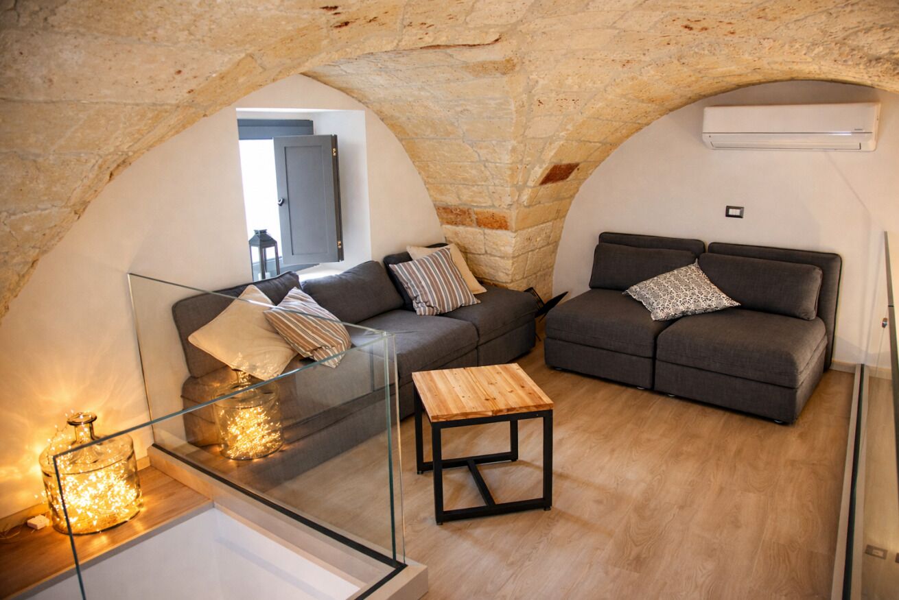 Ein gemütlicher Wohnbereich mit Steinbögen, zwei grauen Sofas mit gemusterten Kissen, einem Glastisch und einem Holznachttisch.