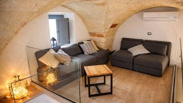 Ein gemütlicher Wohnbereich mit Steinbögen, zwei grauen Sofas mit gemusterten Kissen, einem Glastisch und einem Holznachttisch.