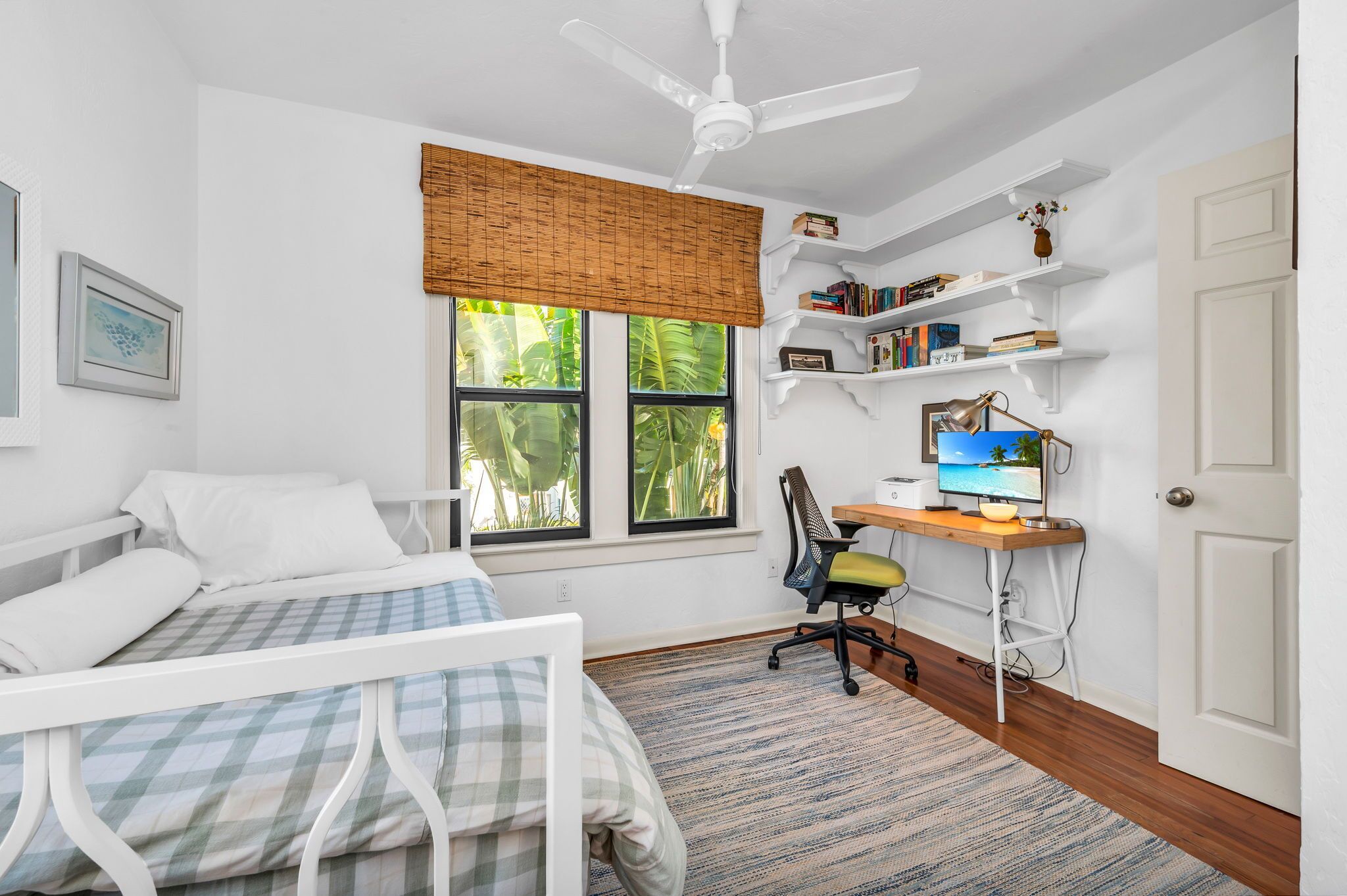 Un dormitorio con cama, escritorio con computadora, silla, ventana con persianas y ventilador de techo.