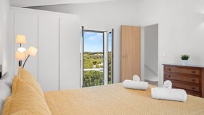 Ein Schlafzimmer mit Bett, Schminktisch, Nachttisch und Lampe. Ein Fenster bietet Blick auf grüne Natur.
