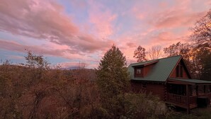 Eine rustikale Holzhütte mit einem grünen Dach, umgeben von dichtem Wald und einem klaren Himmel mit rosa Wolken.