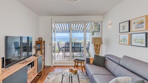A modern living room with a grey sofa, a TV on a wooden cabinet, a small coffee table, and a view of the sea through a glass door. - Sun Spalato Beach & Palms (Split)