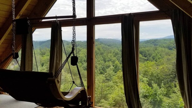 A room with a large window overlooking a forested landscape, a wooden ceiling, and a hanging chair.