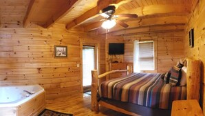 A log cabin bedroom with a large bed, a wooden ceiling fan, a ceiling-mounted TV, and a wooden bathtub.