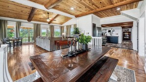 Un'ampia zona giorno con soffitto in legno, un grande tavolo da pranzo in legno e angolo cottura con mobili bianchi e piano di lavoro nero.