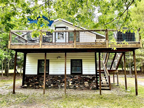 A two-story wooden structure with a balcony, stone foundation, and a house in the background. - River Rock Cabin - Private Escape - ON the Etowah River! Private decks/firepits! (Dahlonega)