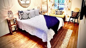 A bedroom with a bed, a nightstand, a chair, a window with curtains, and a wall-mounted lamp.