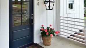 A white house with a dark blue front door, a number '334' above the door, a black lantern on the wall, and a basket of pink flowers on the porch. - Lake Life Retreat 3BR Home w/ 2 Kitchens & Docks (Warwick)