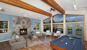 Un vaste salon doté d’une cheminée, d’une table de billard et de grandes fenêtres offrant une vue sur l’extérieur.
