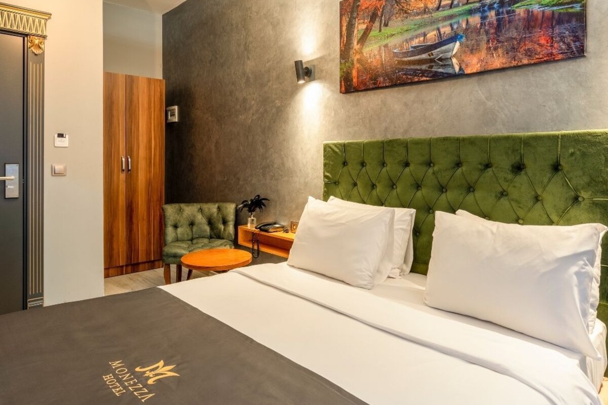 A hotel room with a large bed, a green upholstered headboard, a painting of a landscape, a wooden wardrobe, and a small table with a lamp.