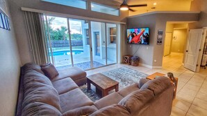 Sala de estar con un televisor grande, un sofá, una mesa de centro y vista a una piscina a través de puertas de vidrio.