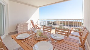 A wooden table set for dining with white plates and glasses, a woven placemat, and a bouquet of white flowers. There are wooden chairs around the table, and a balcony with a railing and a view of buildings. - Global Vacacional: Apartment with sea views, Canet d'en Berenguer (Canet d´en berenguer)