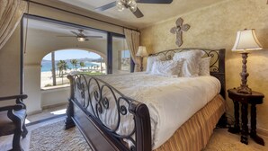 Quarto com cama, ventilador de teto, cruz na parede e vista para a praia.