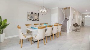 Comedor con mesa y sillas blancas, una araña y una escalera que lleva a otra habitación.
