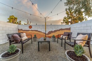 Uma área externa com mesas e cadeiras, cercada por plantas em vasos e um mural decorativo.