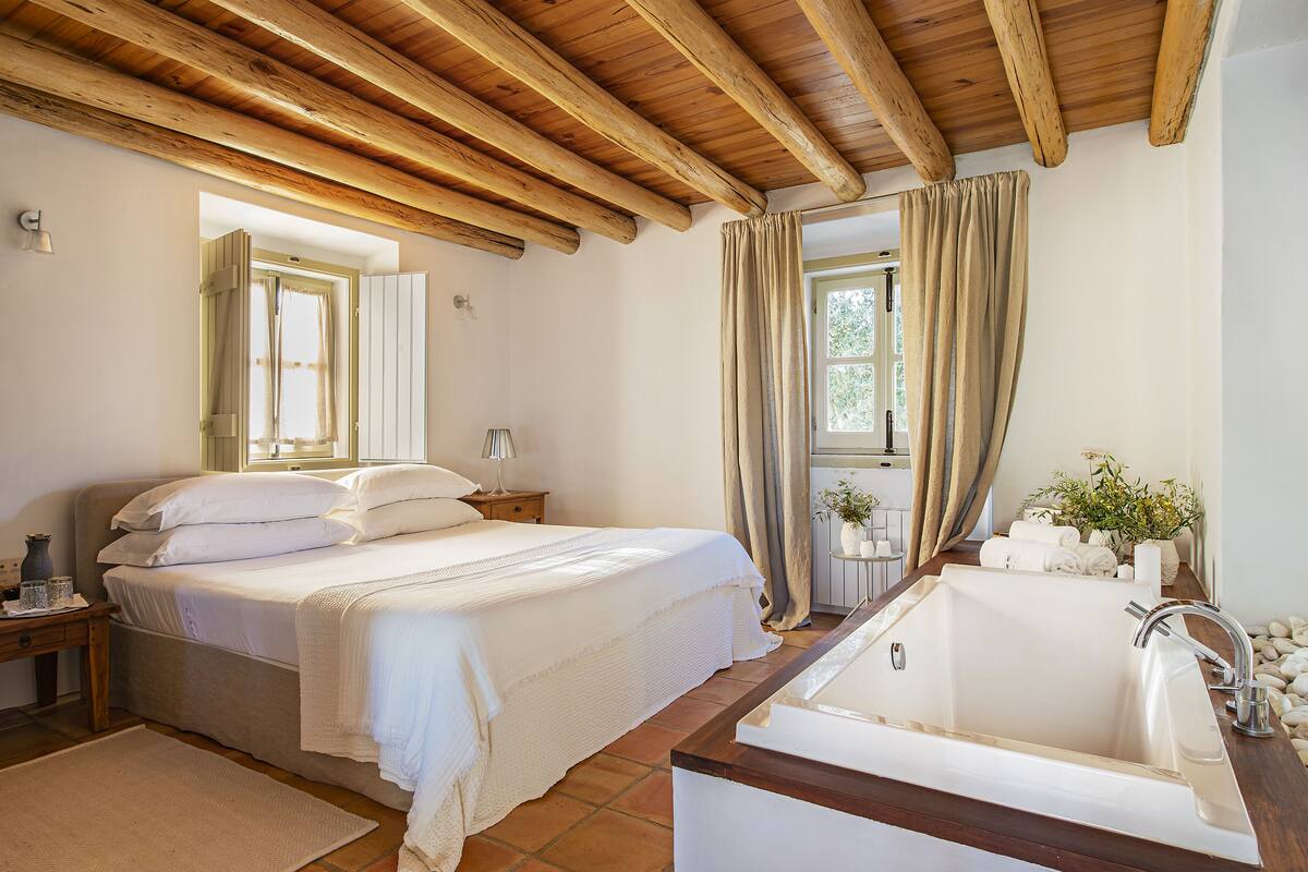 Ein Schlafzimmer mit einem großen Bett, Holzboden, einem Badezimmer mit Badewanne und einem Fenster mit Vorhängen.