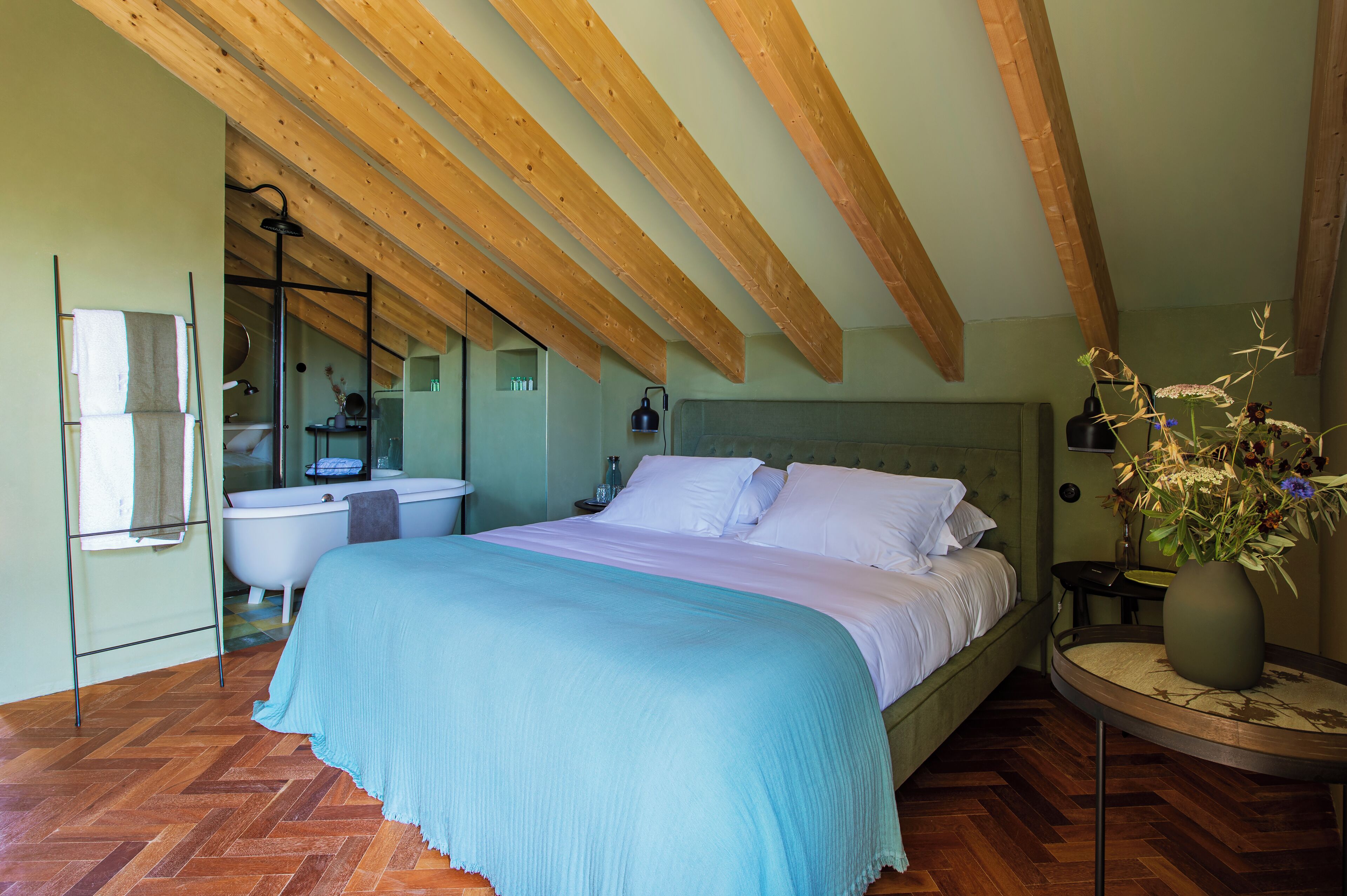 A bedroom with a large bed, wooden beams, a bathroom with a bathtub, and a vase with flowers on a table.