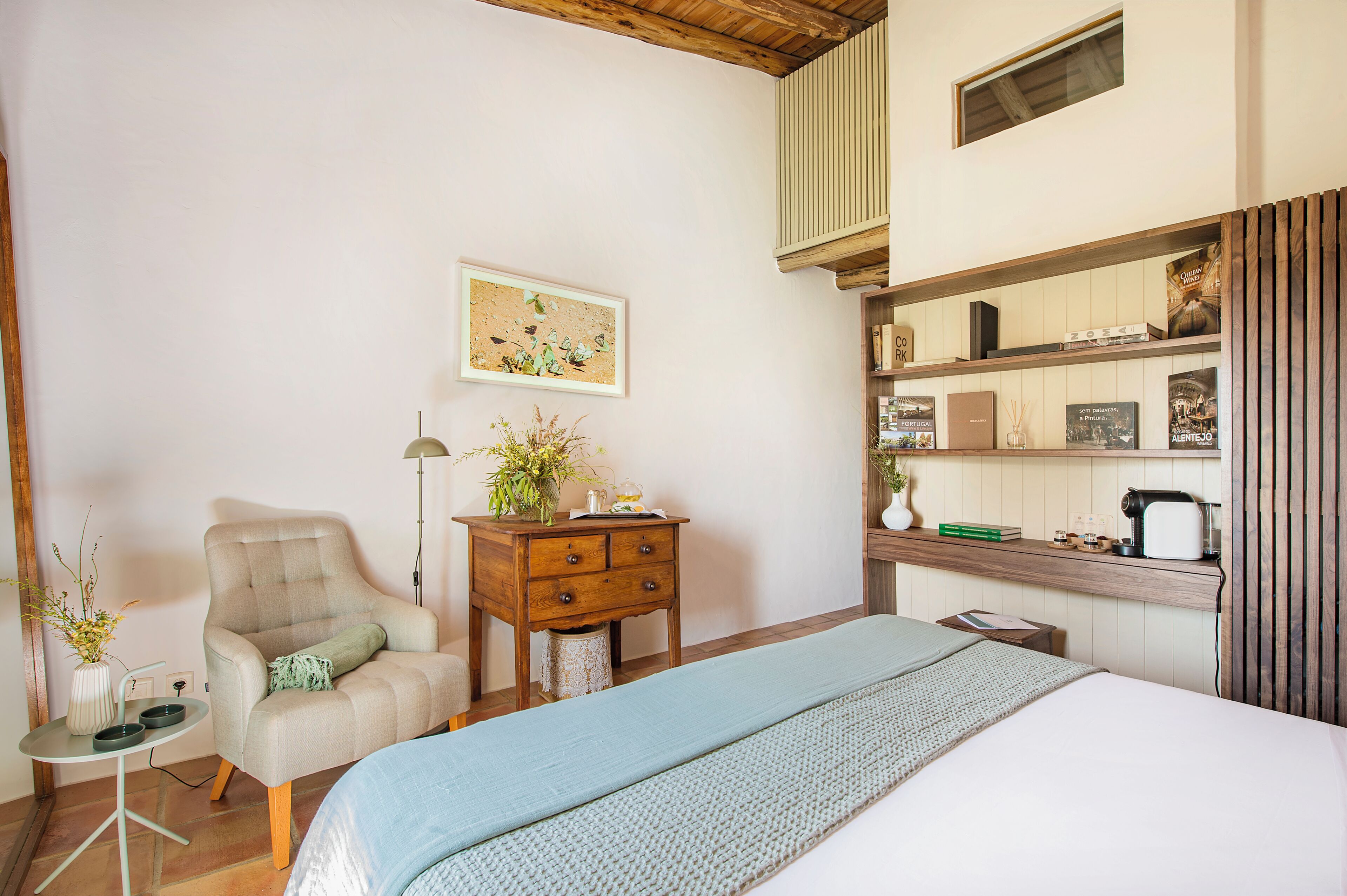 A bedroom with a bed, a chair, a small table, a dresser, and a shelf with various items.
