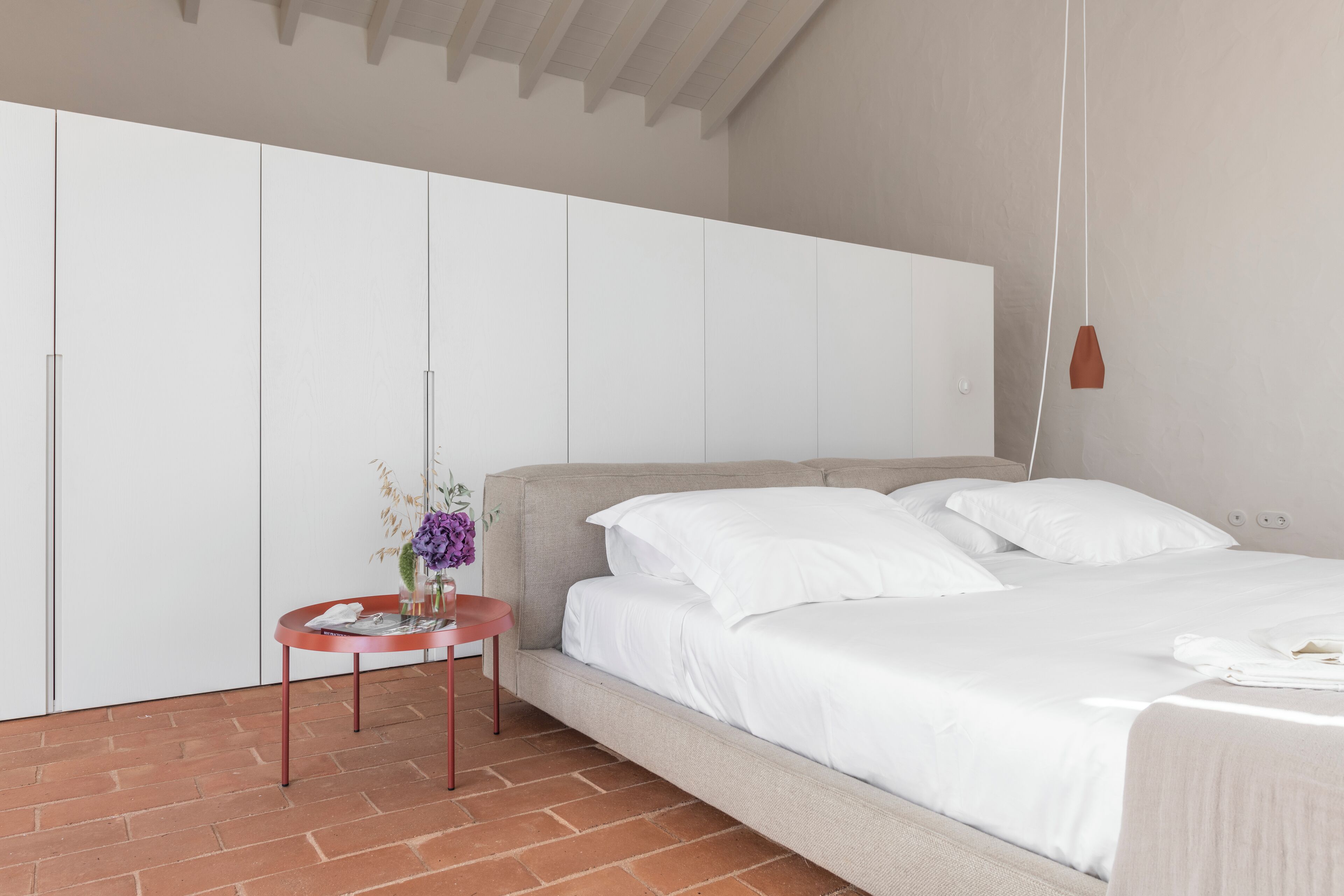 Un dormitorio moderno con una cama grande, una mesita redonda con un florero y un armario blanco.