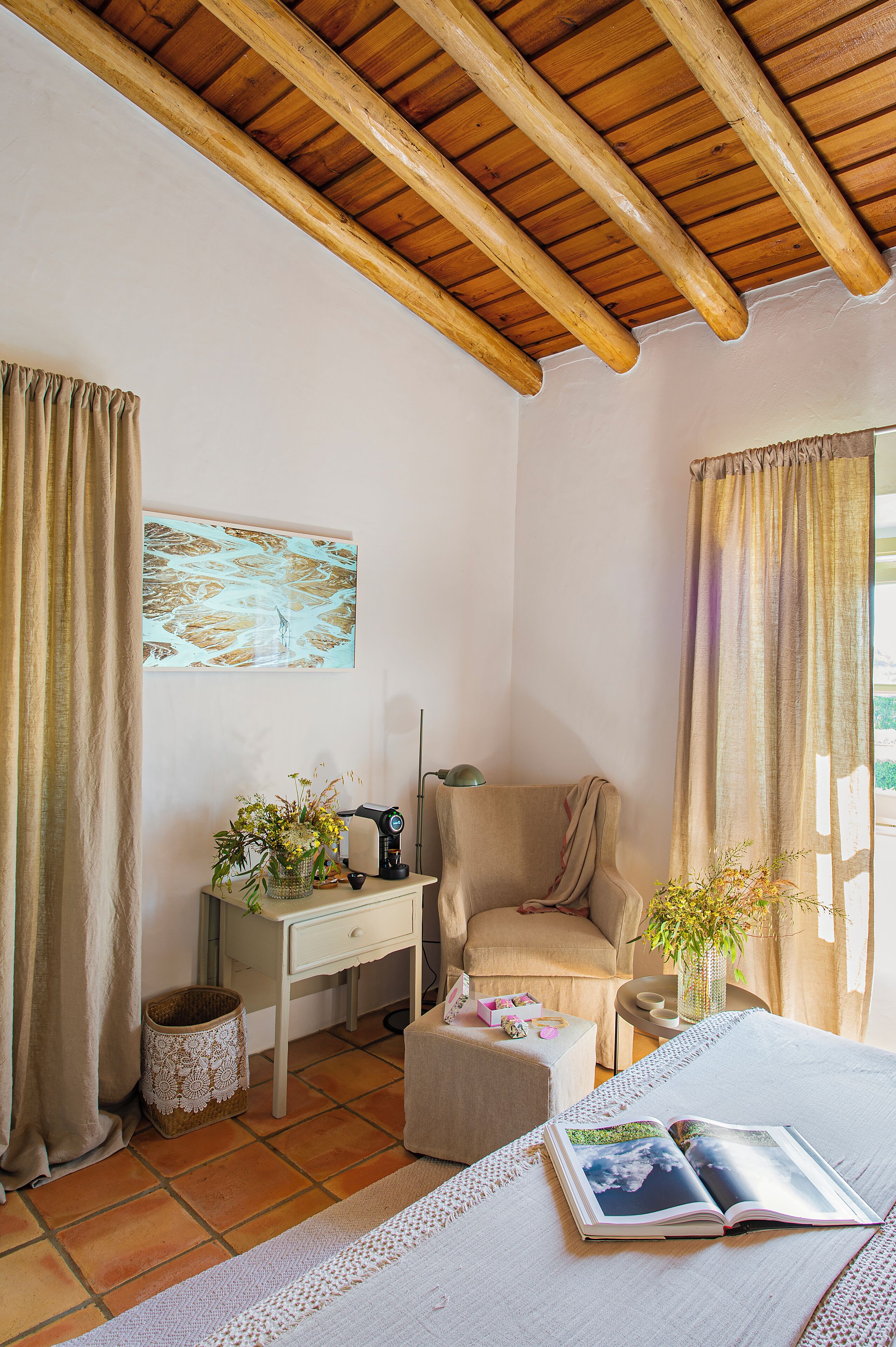 A cozy bedroom with a wooden ceiling, a desk with a lamp, a chair, a bedside table with a plant, and a bed with a book.