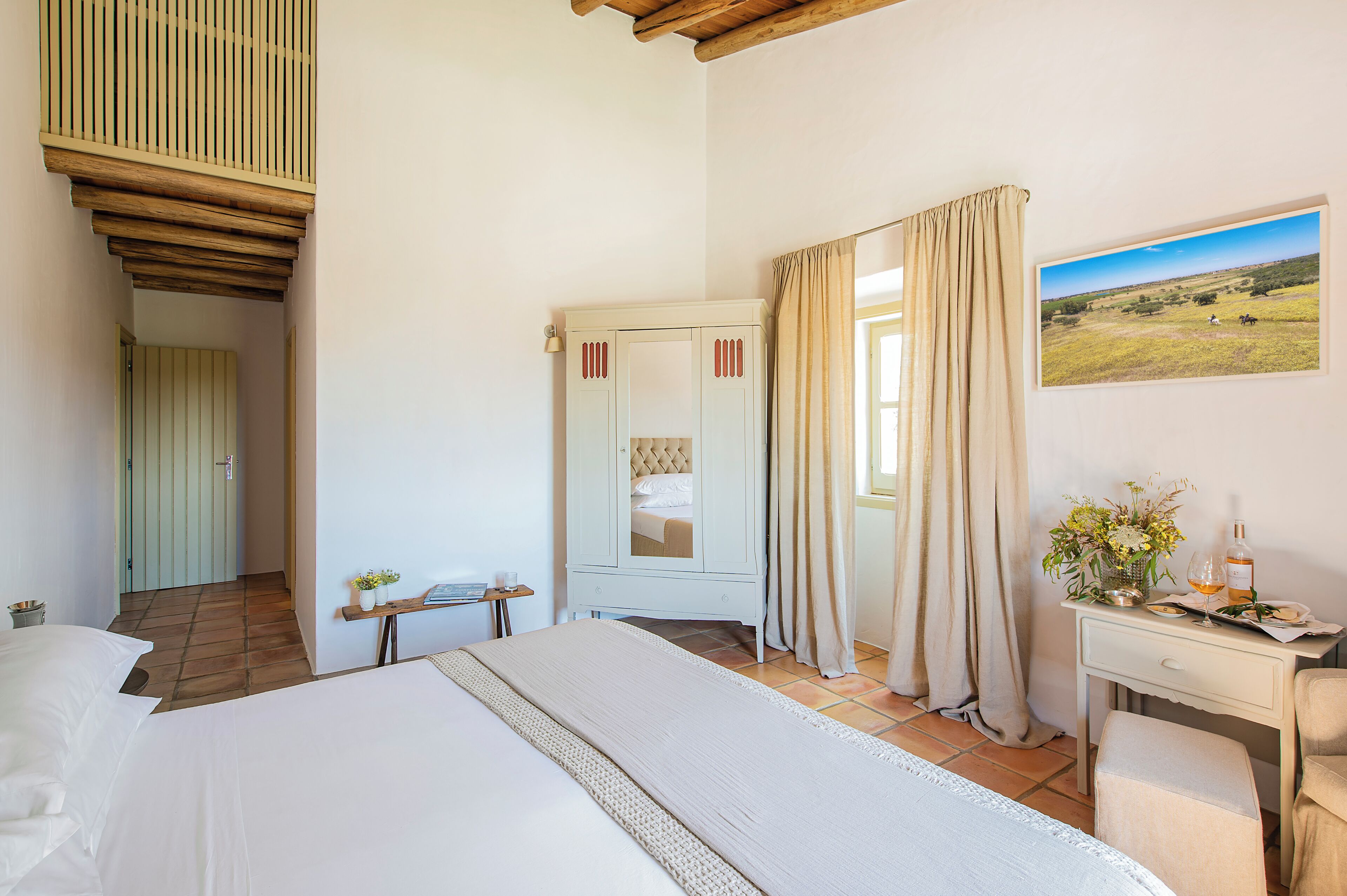 Une chambre à coucher avec un grand lit, un bureau agrémenté d’un vase de fleurs et un tableau représentant un paysage.