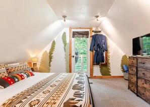 A bedroom with a sloped ceiling, a bed with patterned bedding, a dresser, a television, and a window with a view of trees. - Romantic Cabin: Hot Tub, Fire Pit, Indoor & Outdoor Living, EV Charger (Martin)