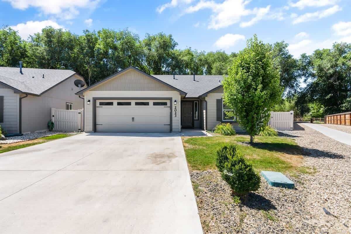 A two-story house with a garage, a small front yard with a tree, and a gravel driveway.