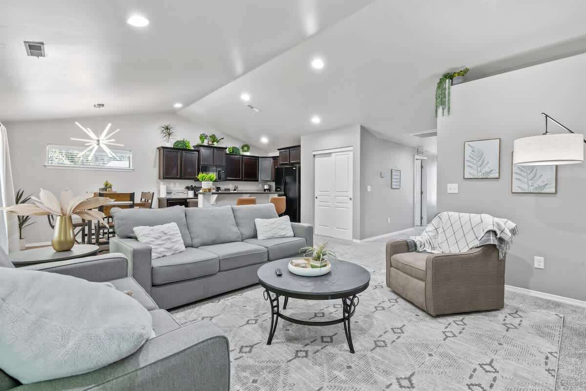 A modern living room with a grey sofa, a coffee table, and a grey armchair. There is a kitchen in the background with dark cabinets and a refrigerator.