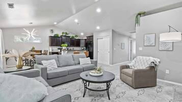A modern living room with a grey sofa, a coffee table, and a grey armchair. There is a kitchen in the background with dark cabinets and a refrigerator.