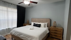 Un dormitorio con cama, ventilador de techo, mesita de noche, lámpara y ventana con cortinas.
