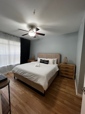 A bedroom with a bed, a ceiling fan, a bedside table, a lamp, and a window with curtains.