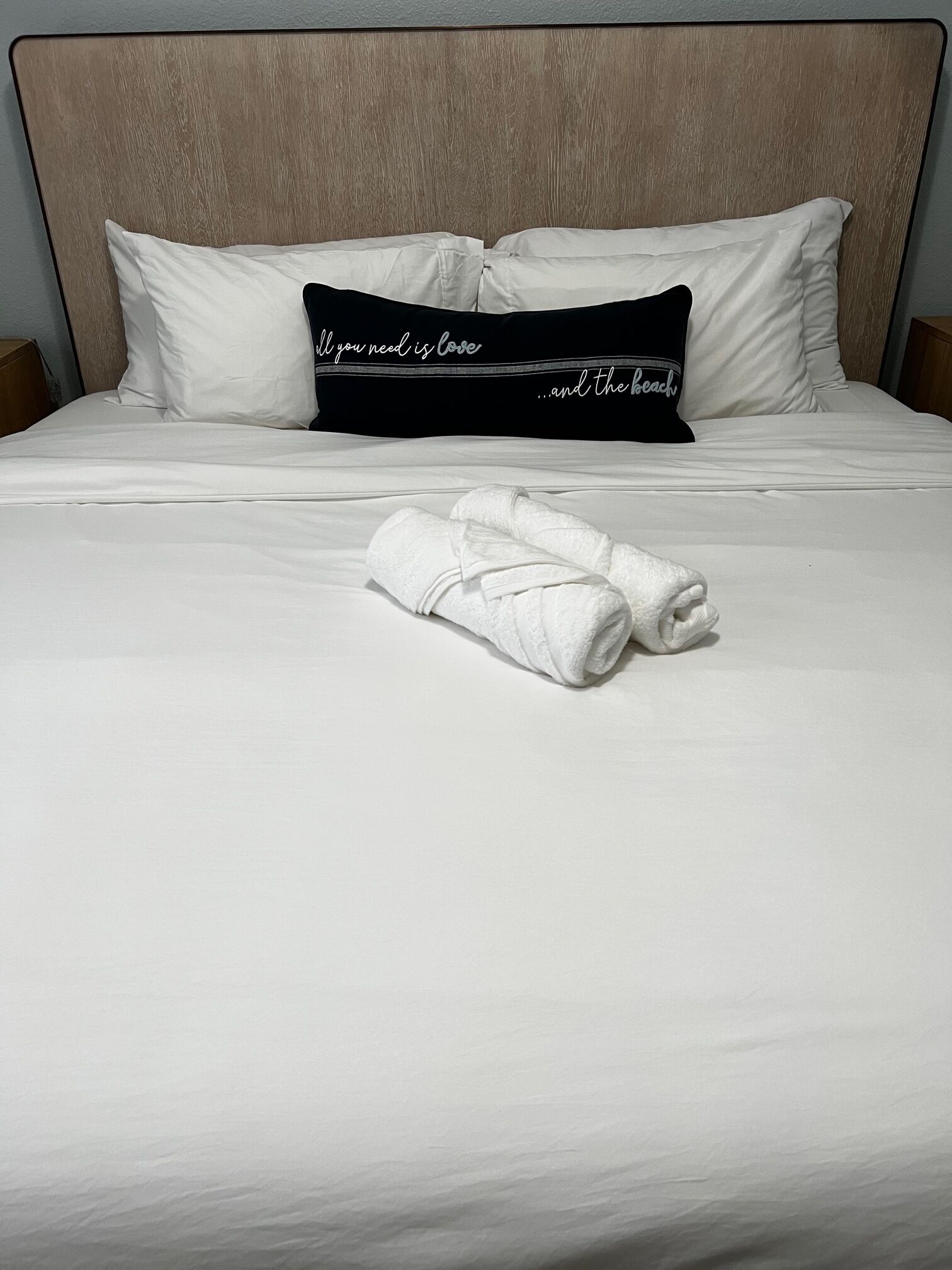Un lit bien fait, avec des draps blancs, un oreiller noir orné de lettres blanches et deux serviettes enroulées.
