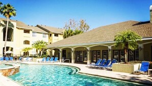 Una piscina con sillones de descanso y palmeras, rodeada por un edificio con techo de tejas y varias ventanas.