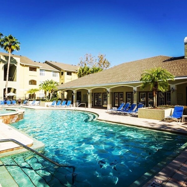 A swimming pool with lounge chairs and palm trees, surrounded by a building with a tiled roof and multiple windows.