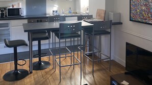 A modern kitchen with white cabinets, a stainless steel range hood, and a built-in microwave. There are three black bar stools and a wooden dining table. A colorful abstract painting is on the wall, and a flat-screen TV is on a wooden stand.