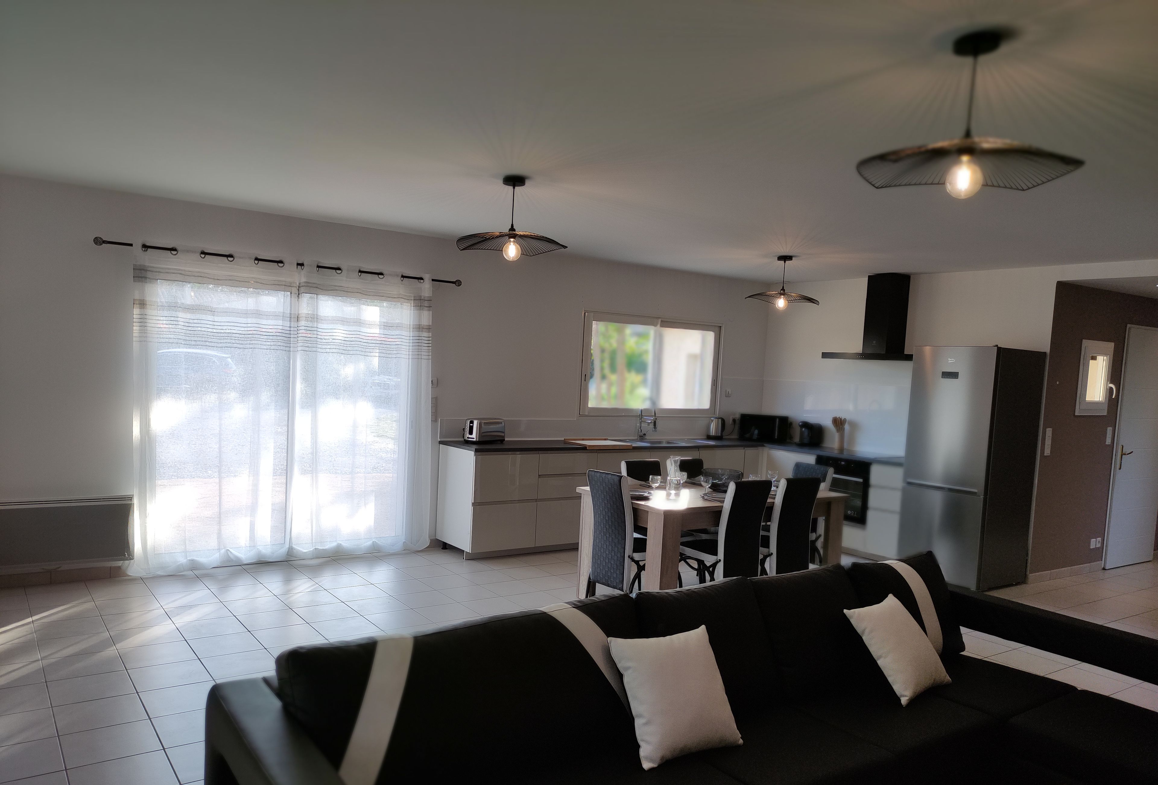 A modern living room with a black sofa, dining table, and chairs. There is a kitchen area with a refrigerator, microwave, and stove. The room has large windows with sheer curtains.