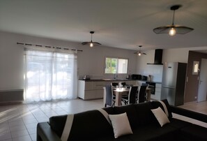 A modern living room with a black sofa, dining table, and chairs. There is a kitchen area with a refrigerator, microwave, and stove. The room has large windows with sheer curtains. - House with swimming pool for rent from Sunday to Sunday (Labeaume)