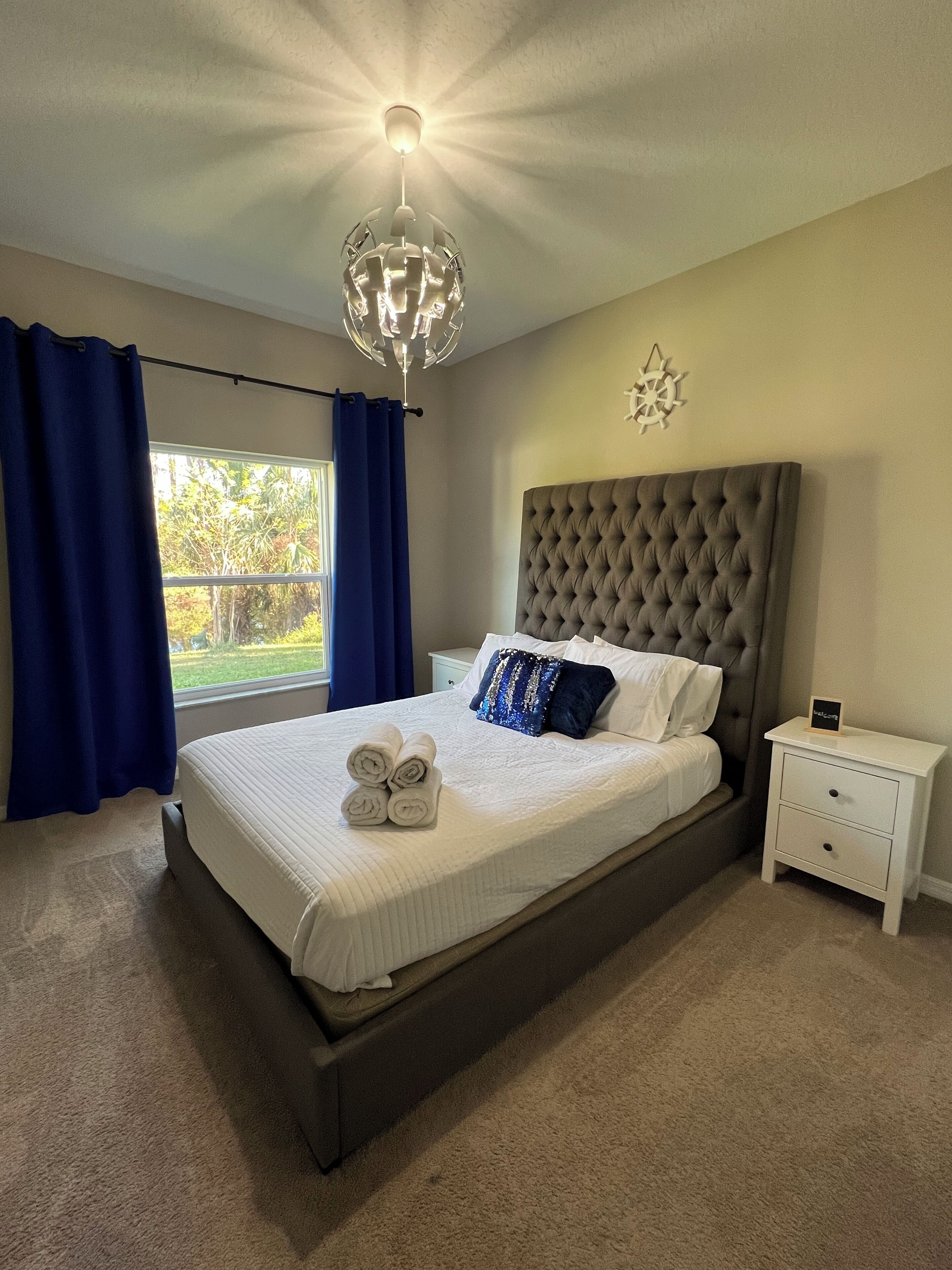A bedroom with a large bed, a nightstand, a chandelier, and a view of greenery outside.