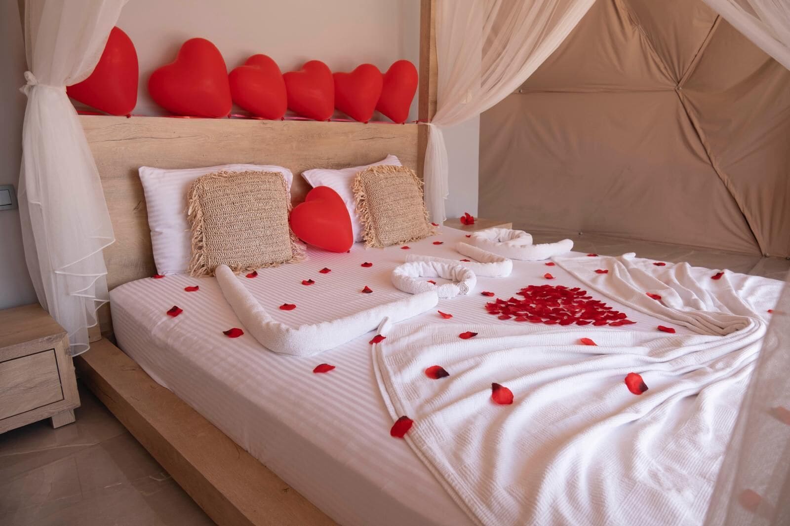 A neatly made bed with rose petals and a heart-shaped decoration, a wooden headboard, and a white nightstand.