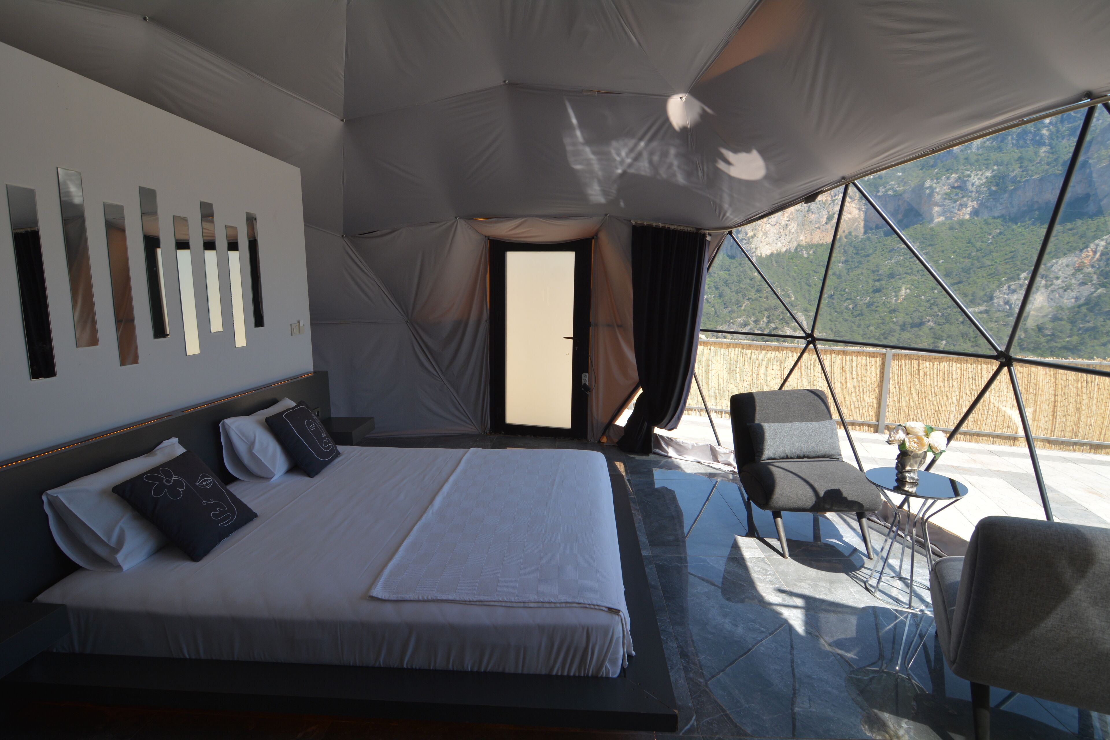 Quarto com cama, almofadas, mesa de cabeceira e vista para uma paisagem montanhosa por grandes janelas.