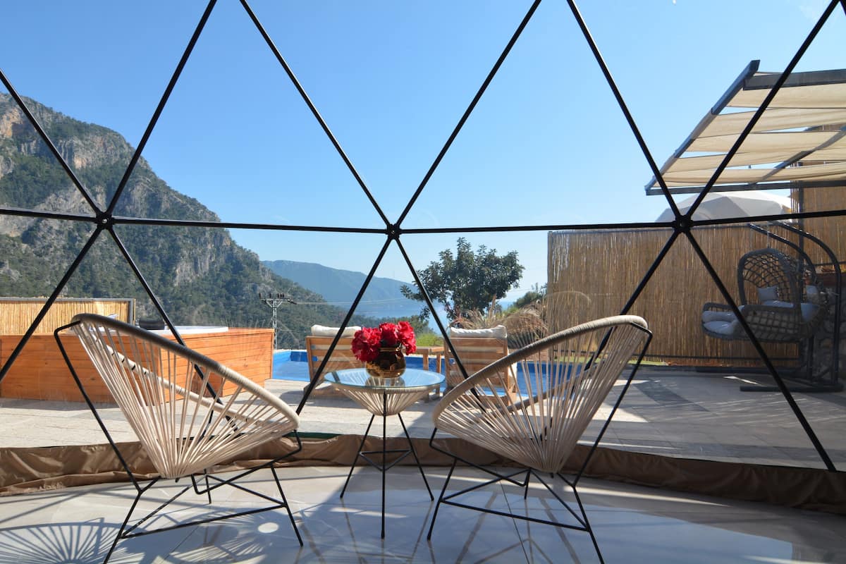 A terrace with wicker chairs, a table with a vase of red flowers, and a view of mountains.