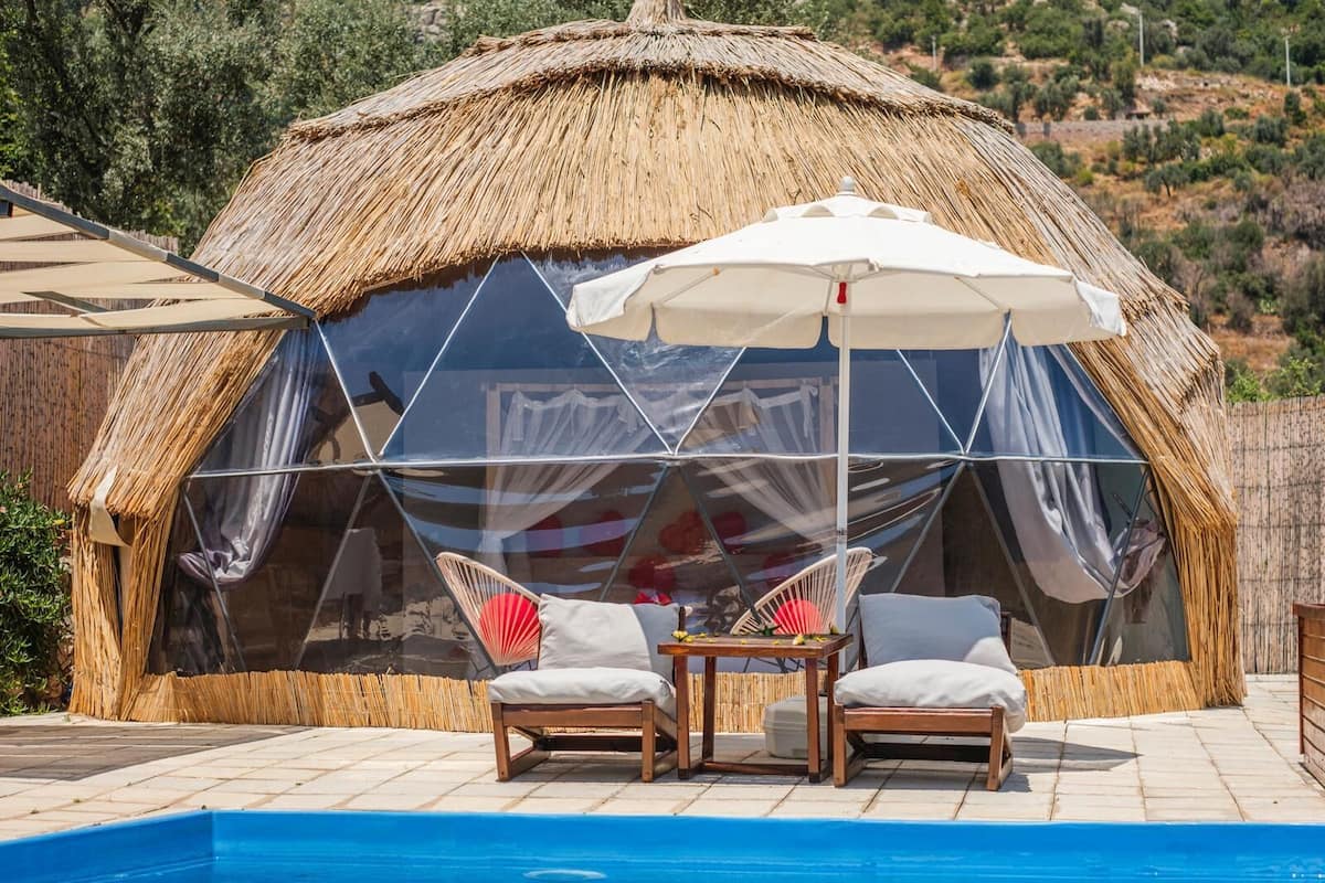 A thatched-roof structure with a glass enclosure, two wooden chairs, and a table with umbrellas.